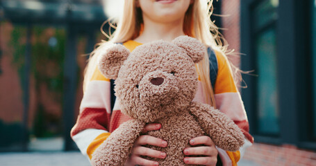 Standing in front of camera and holding teddy bear with both hands. Holding backpack on her back.