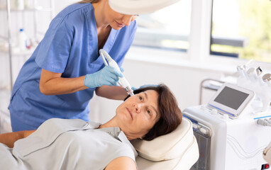 Elderly woman getting facial deep hydration procedure from experienced cosmetologist in aesthetic...