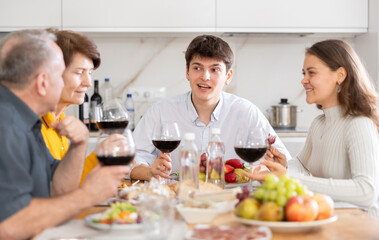 Young grown son with wife and parents having cheerful conversation at family holiday table in light kitchen