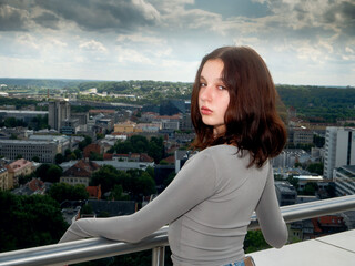 Young teenager in a casual blue jeans and grey shirt standing on a balcony, beautiful city in a background Kaunas town, Lithuania. Travel and tourism concept. Model has slim body type.