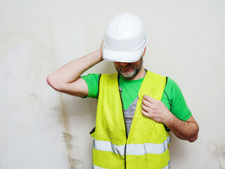 Constriction worker in green t shirt and grey rough overalls and white plastic safety helmet and reflective vest on light color background. The model is in his 50s with grey beard.