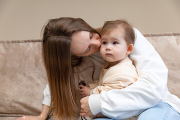 Young caring mother hugged her little daughter, pressing her to her chest. Woman holding a newborn baby in her arms on a sofa at home. Smiling mother hugging a small newborn baby showing love