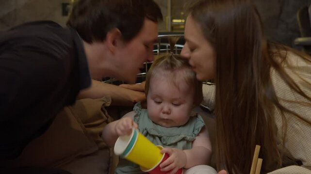 Mother and father are kissing their child on both sides, while the daughter is excitedly playing with colorful cups, preparing for her upcoming birthday.