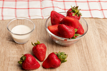 desayuno saludable, fresas en un bol de vidrio, frutillas sobre la mesa, un vaso con crema sobre una mesa de madera, al fondo un paño de cocina blanco con líneas rojas