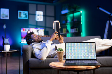 Man lounged on cozy couch reading ebooks on tablet next to laptop with stock market financial...
