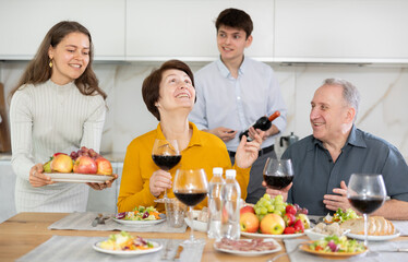 Mother and father visiting young couple, sitting with glass of wine in kitchen. Happy girl serving fruit, excited guy in background