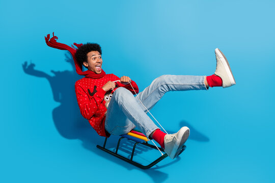 Full size photo of pretty young man ride sledge fluttering scarf wear red christmas sweater masquerade theme party on blue color background