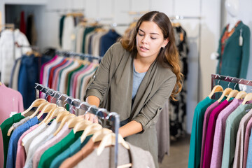 Positive young woman choosing season outfit clothes among the racks in a shopping mall. High quality photo