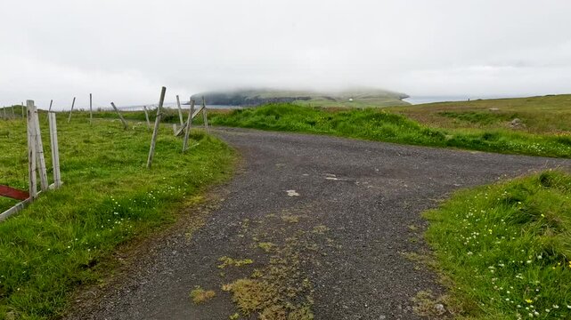 Moving forward on a gravel path with rainfall. Low clouds hang over the remote Scandinavian island of N&oacute;lsoy in the Faroe Islands. Dramatic landscape of grass covered mountains and basalt columns.