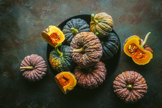 Variety of green and orange heirloom pumpkins, some cut open exposing orange flesh, arranged in black bowl and on custom teal and copper background
