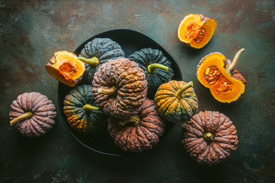 Variety of green and orange heirloom pumpkins, some cut open exposing orange flesh, arranged in black bowl and on custom teal and copper background