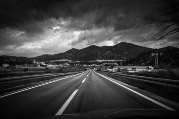 Monochrome Rainy Road with Speed and Danger Leading to Mountains Under Dramatic Skies