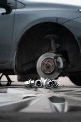 In the foreground, loose bolts and the removed wheel rest on the ground, while in the background, a car lifted on a jack reveals its exposed brake disc and suspension system.