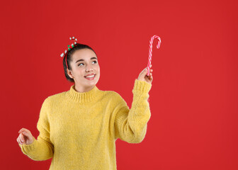 Young woman in yellow sweater and festive headband holding candy cane on red background, space for text