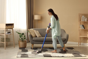 Smiling young woman cleaning rug with cordless vacuum cleaner in living room