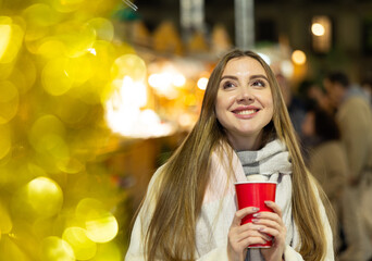 Cute happy woman with paper cup of hot drink walking at christmas fair