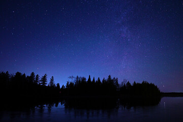 A night scene of a star filled sky and a faint glow of the Milky Way over a calm fresh water lake