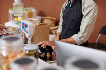 Selective focus on shopkeeper measuring two freshly harvested eggplants on modern weigh scale. Male vendor checking the weight and price of organic vegetables at cashier desk.