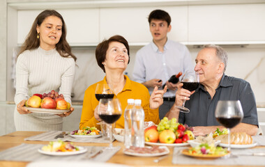 Mother and father visiting young couple, sitting with glass of wine in kitchen. Happy girl serving fruit, excited guy in background