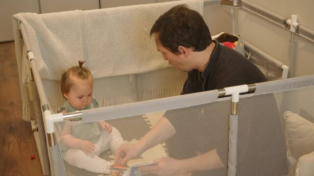 Young father is teaching his daughter how to read with help of children's book, while they are sitting in playpen designed for young children.