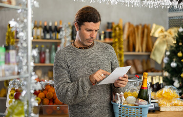 Portrait of modern man with cart shopping at big supermarket, looking for something and reviewing and checking list of products to