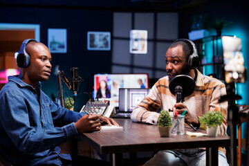 African American cohosts engaged in a lively discussion, recording podcast in home studio equipped with professional audio gadgets and RGB lights. Black men filming show with cell phone on tripod.