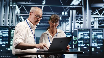 Old employees looking around high tech data center, using laptop to crosscheck disaster recovery plan and assess server electronics in need of replacement, preventing crashes