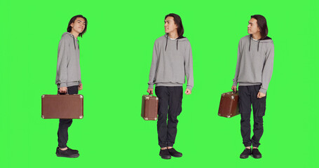 Person waiting impatiently in studio, looking around and checking hour on wristwatch. Young adult carrying retro briefcase standing over full body greenscreen backdrop, acting impatient.