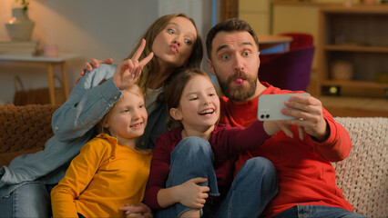 Fototapeta premium Caucasian happy family mother father kids little girls daughters posing grimace photo on mobile phone at evening home funny parents children record video online blog smartphone selfie social media app