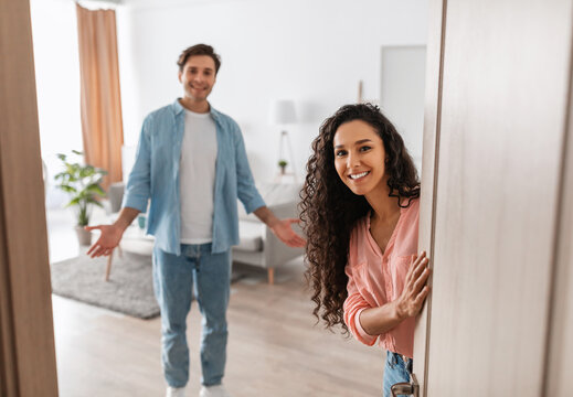 Portrait of cheerful couple inviting guests to enter home, happy people standing in doorway of modern flat, looking out waiting for visitor to come in, receiving friend, lady peeking out of front door