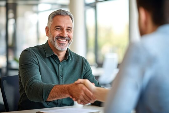 Middle aged man shaking hands with younger partner in office meeting for business collaboration agreement