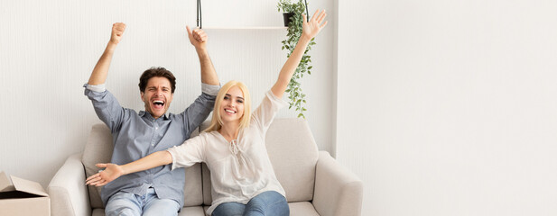 A cheerful couple sits on a sofa with their arms raised in excitement, sharing a joyful moment....