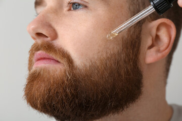 Young bearded man applying essential oil onto his face on grey background, closeup
