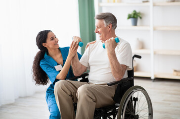 Millennial physiotherapist helping disabled elderly man in wheelchair to work out with dumbbells,...