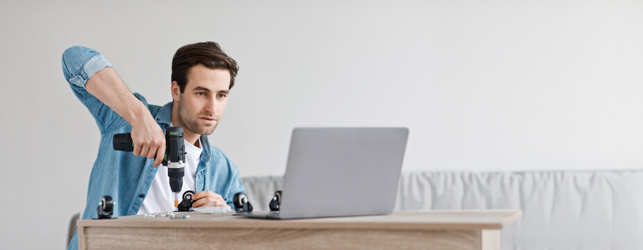 Online lesson and instructions for assembling furniture with own hands. Concentrated young handsome man collects table with electric screwdriver and looks at laptop in living room, empty space