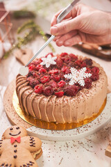 A close-up of a festive chocolate cake being carefully decorated by hand. The cake is topped with fresh raspberries, blackberries, grated chocolate, and intricate white snowflake decorations