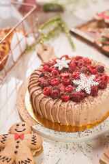 A close-up of a festive chocolate cake being carefully decorated by hand. The cake is topped with fresh raspberries, blackberries, grated chocolate, and intricate white snowflake decorations