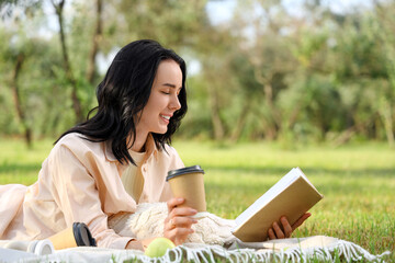 Pretty young woman with cup of coffee reading book on blanket in park