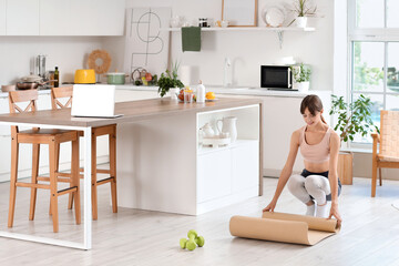 Sporty young woman rolling out fitness mat in kitchen