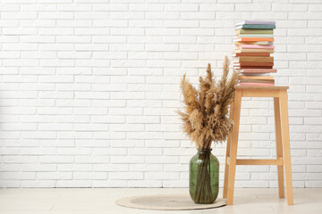 Wooden stool with stack of books and pampas grass in vase near white brick wall