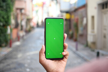 A Hand Firmly Holding a Smartphone with a Green Screen Display in a Vibrant Urban Alley