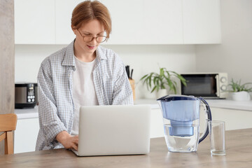Young man with water filter jug using laptop on table in kitchen