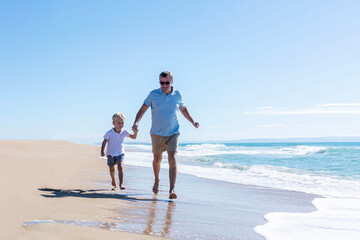 Little boy with an elderly man running along the sandy seashore on a sunny day. The child is wearing a white T-shirt and gray shorts. Love and tenderness of a grandfather with his grandson.