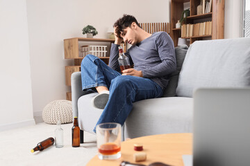 Drunk young man with whiskey sitting on sofa at home. Alcoholism concept