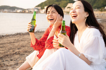 Obraz premium Two Women Relaxing on the Beach with Beers at Sunset