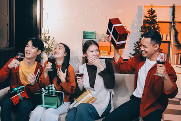 Group of young Asian man and women as friends having fun at a New Year's celebration, holding gift boxes standing by Christmas tree decoration, midnight countdown Party at home with holiday season.