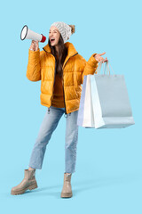 Young woman in winter clothes with shopping bags shouting into megaphone on blue background