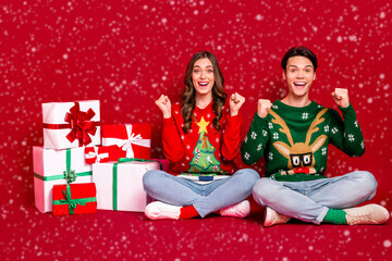 Full length photo of two delighted astonished people sit floor raise fists pile stack newyear giftbox isolated on red color background