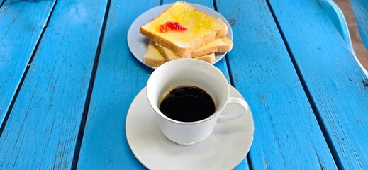 Hot coffee served in white glass with buttered bread and strawberry jam on a bright blue wooden table. Espresso in a glass for morning drink. Three slices of buttered toast served with strawberry jam.