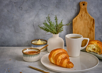 A fresh croissant with a crisp on a white plate with a cup of black coffee on a gray background.